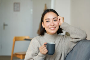 Smiling asian woman sitting at home with cup of coffee or tea