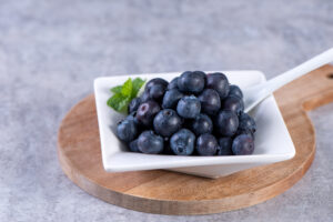 Pile of blueberry fruit in a bowl plate on a tray over gray cement concrete background, close up, healthy eating design concept.
